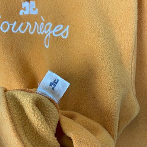 Puede incluir: Primer plano de una prenda de forro polar amarillo mostaza. La palabra "Courr&egrave;ges" est&aacute; escrita en escritura blanca en la parte superior. Se ve una peque&ntilde;a etiqueta con un logotipo. La tela tiene un aspecto suave y texturizado.
