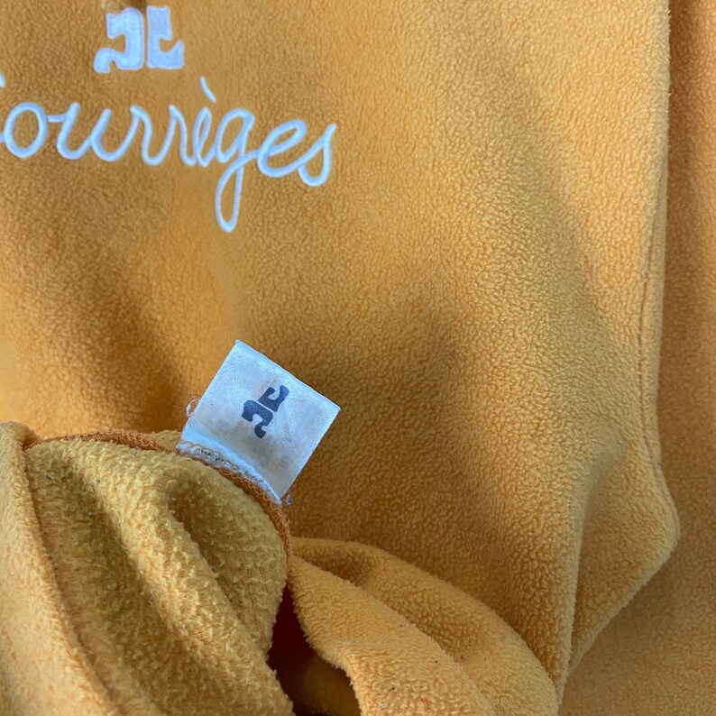 Puede incluir: Primer plano de una prenda de forro polar amarillo mostaza. La palabra "Courr&egrave;ges" est&aacute; escrita en escritura blanca en la parte superior. Se ve una peque&ntilde;a etiqueta con un logotipo. La tela tiene un aspecto suave y texturizado.