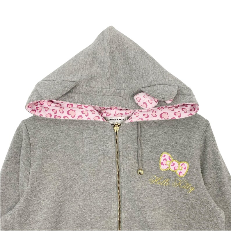 May include: A gray zip-up hoodie with a hood featuring cat ears and a pink leopard print lining. The hoodie has a gold zipper and a Hello Kitty bow applique with gold embroidery. The Hello Kitty logo is embroidered below the bow.