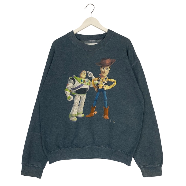 Op de afbeelding: Een donkergroen sweatshirt met een afbeelding van Buzz Lightyear en Woody uit Toy Story. Buzz draagt zijn witte, groene en paarse ruimtepak, terwijl Woody een cowboyhoed en vest draagt. Het sweatshirt hangt aan een witte hanger.
