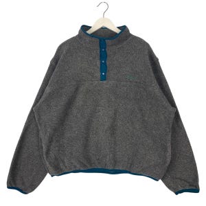 Könnte beinhalten: Ein grauer Fleece-Pullover mit türkisfarbenem Kragen, Knopfleiste und Besatz. Das langärmlige Hemd hängt an einem weißen Kleiderbügel. Ein kleines Logo ist auf der Brust sichtbar.