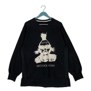 Op de afbeelding: Zwart sweatshirt met een witte grafische print van een cartoonfiguur met een mohawk en een bril. De tekst "Devil" is gedrukt op het voorhoofd van het figuur. De tekst "DEVILOCK NIGHT" is gedrukt onder het figuur.