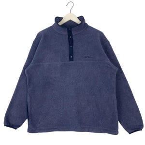 Puede incluir: Un jersey de forro polar azul con cuello alto y tres botones azul marino. La camisa de manga larga tiene un pequeño logotipo bordado en el pecho y está colgada de una percha blanca. El material de forro polar parece suave y cálido.