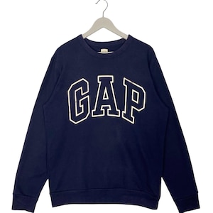 May include: A navy blue sweatshirt featuring the word "GAP" in large, white, outlined letters. The long-sleeved top has a crew neck and is suspended from a metal hanger. The garment is a classic style.