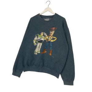 Op de afbeelding: Donker teal sweatshirt met een afbeelding van Buzz Lightyear en Woody uit Toy Story. De personages zijn in kleur, met Buzz in zijn witte en groene ruimtepak en Woody in zijn cowboy-outfit. Het sweatshirt hangt aan een witte hanger.