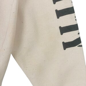 May include: Close-up of a cream-colored sweatshirt sleeve. The sleeve features a dark gray, stylized text design. The fabric has a slightly textured appearance. The design is partially visible, with the letters appearing to be bold and blocky.