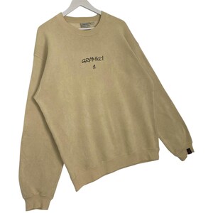 May include: A tan Gramicci sweatshirt with a crew neck and long sleeves. The brand name and logo are printed in black on the chest. The sweatshirt has ribbed cuffs and hem. The garment is hanging on a white hanger.