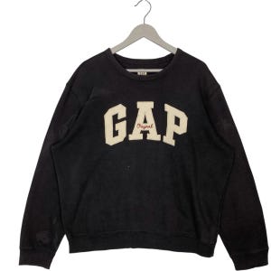 May include: Black crew neck sweatshirt with the word "GAP" in large, off-white, block letters across the chest. The word "Original" is printed in a smaller, red font below the "GAP" logo. The sweatshirt is hanging on a grey hanger.