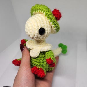 Könnte beinhalten: Handgemachte Häkelpuppe mit cremefarbenem Kopf, grünem Körper und roten Akzenten. Die Puppe hat einen grünen Hut mit rotem Bommel und schwarzen Knopfaugen. Die Puppe wird in einer Hand gehalten.