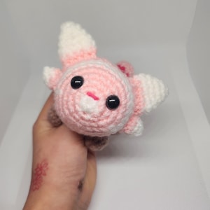 May include: A pink and white crocheted plush toy with black eyes and a pink nose. The toy is in the shape of a small animal with large ears.
