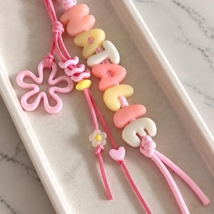 May include: A personalised keychain featuring pink, yellow, and white letter beads spelling the name NATALIE on a pink cord. The accessory includes a pink flower-shaped charm, a small yellow bead, a tiny white flower bead, and a small pink heart bead.