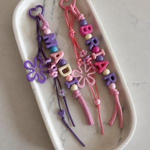 May include: Two beaded keychains featuring heart-shaped clasps and cord loops. One purple keychain spells MADDI with a flower charm, while the pink one spells BRIAR with a flower charm. Both include various round, letter, and shaped beads.