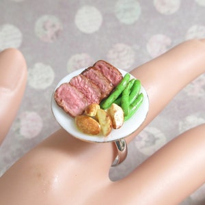May include: A silver ring with a miniature white plate featuring a slice of roasted meat, green beans, and potatoes.