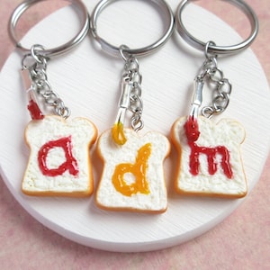 Puede incluir: Tres llaveros con forma de rebanadas de pan con diferentes letras escritas en jalea. Las letras son "a", "d" y "m". La jalea es roja y amarilla.