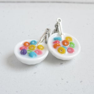 Op de afbeelding: Een paar novelty oorbellen met miniatuur witte kommen gevuld met kleurrijke ontbijtgranen en melk. Elke oorbel heeft een kleine zilveren lepel en een zilveren haakje. De ontbijtgranen zijn in verschillende kleuren.