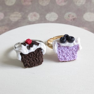 Pode incluir: Dois anéis de cupcake, um com cobertura de chocolate e uma framboesa vermelha, o outro com cobertura roxa e mirtilos.