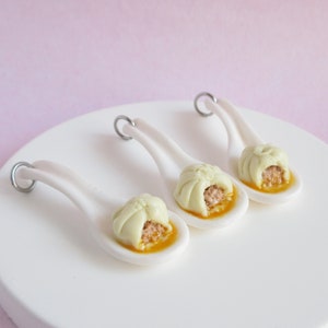 May include: Three white ceramic spoons with a small, open-faced dumpling on each. The dumplings are filled with a brown filling and have a yellow sauce around them.