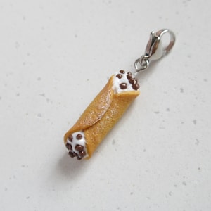 Könnte beinhalten: Ein Miniatur-Cannoli-Anhänger mit einem silbernen Verschluss. Der Cannoli ist mit weißer Creme gefüllt und mit Schokoladenstückchen bestreut.
