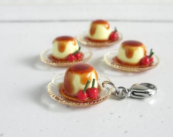 Puede incluir: Cuatro postres de crema de caramelo en miniatura con cerezas rojas encima en platos de vidrio transparente. Los postres están unidos a un dije plateado.