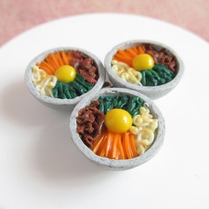 Puede incluir: Tres cuencos en miniatura de bibimbap, un plato coreano, hechos de arcilla. Cada cuenco contiene arroz, verduras, carne y un huevo frito.