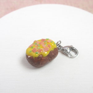 May include: A small, brown, baked potato charm with a yellow cheese topping, pink ham pieces, and green chives. The charm is attached to a silver lobster clasp.