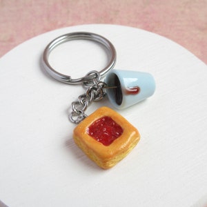 以下が含まれることがあります： 小さな青いセラミックカップに茶色の液体が満たされ、赤いゼリーフィリングの入った四角いペストリーが付いたシルバーキーチェーン