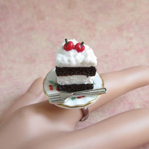 May include: A miniature chocolate cake with white icing and red cherries on top, sitting on a small white plate with a silver fork. The plate is attached to a silver ring.