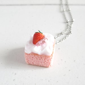 May include: A small pink cake pendant with white icing, decorated with a red strawberry and silver beads. The cake is suspended from a silver chain, forming a quirky necklace.