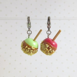 May include: Two miniature caramel apples with a stick, one green and one red, are attached to silver charm clips. The apples are covered in caramel and chopped nuts.