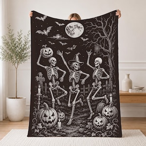 May include: A black and white throw blanket featuring a detailed illustration of three skeletons dancing in a graveyard. The scene includes a full moon, flying bats, a bare tree, carved jack-o'-lanterns, and lit candles under a night sky.