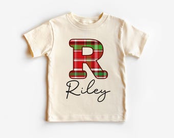 May include: Cream-colored t-shirt featuring a large, red and green plaid letter "R" and the name "Riley" in a cursive font. The shirt is a short-sleeved, crew neck style.