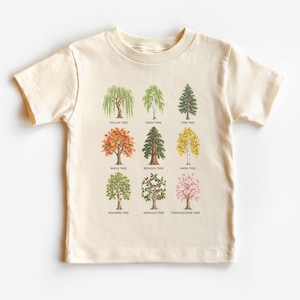 May include: A cream-colored short-sleeve t-shirt featuring a grid of nine illustrated trees. Each tree is labeled with its name: Willow Tree, Birch Tree, Pine Tree, Maple Tree, Redwood Tree, Aspen Tree, Sycamore Tree, Magnolia Tree, and Cherry Blossom Tree.