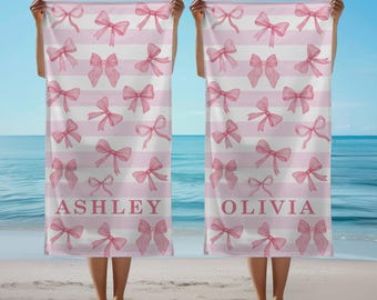 May include: Two beach towels with a pink bow pattern on a white and pink striped background. The word "ASHLEY" is printed on one towel and "OLIVIA" on the other. The towels are held up on a beach.