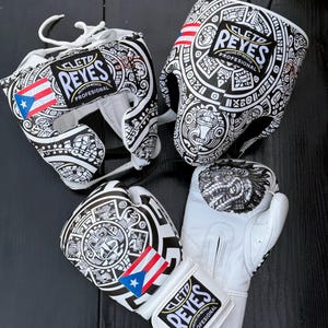 May include: A set of white boxing gear, including a head guard and gloves, with black tribal-style patterns and the text "REYES". The gloves and headgear feature flags of Puerto Rico and the United States.