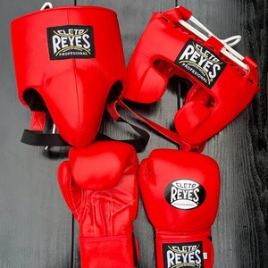 Könnte beinhalten: Ein Set aus leuchtend roter Boxausrüstung, bestehend aus zwei Kopfschützern und zwei Boxhandschuhen. Jeder Artikel trägt ein schwarz-weißes Cleto Reyes Profesional-Etikett. Die Kopfschützer haben weiße Schnürverschlüsse und schwarze Riemen.