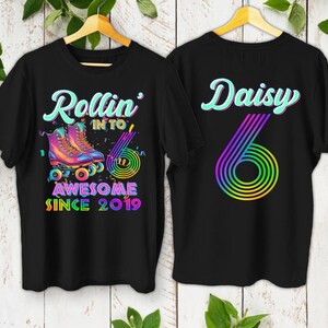 May include: Black t-shirts with roller skate graphics. One shirt says "Rollin' into Awesome Since 2019" with colorful roller skates. The other shirt says "Daisy 6" with a rainbow design. Both shirts are hanging on wooden hangers.