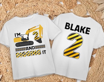 May include: Two white t-shirts with a construction theme. One shirt features a yellow excavator with the text "I'm 2 and digging it". The other shirt has a black number 2 with yellow stripes and the name "Blake" in black text.
