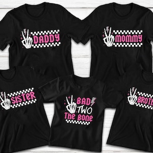 May include: Black t-shirts with matching designs for a family. Each shirt features a skeleton hand making a peace sign, with text like "Daddy," "Mommy," "Sister," "Brother," and "Bad Two The Bone" in pink and white.