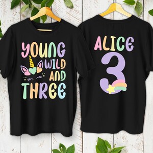 May include: Two black t-shirts with colorful text and graphics. One shirt says "YOUNG WILD AND THREE" with a unicorn graphic. The other shirt says "ALICE 3" with a rainbow and star graphic. Both shirts are hanging on wooden hangers.