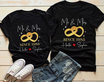 May include: Two black t-shirts with the text "Mr & Mrs" and "SINCE 1985" in white and gold. The design includes two gold interlocking rings and the names "Hunter" and "Sophia" with a heart symbol. The shirts are displayed with white sneakers and denim shorts.