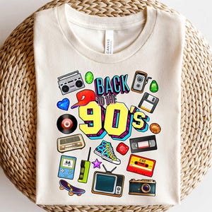 May include: A white t-shirt with a colorful graphic design featuring the text "Back to the 90's" and retro icons like a boombox, a camera, a cassette tape, and a video game console.