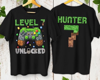 May include: Two black t-shirts with gaming designs. One has "LEVEL 7 UNLOCKED" and a pixelated controller. The other shows "HUNTER 7" with a pixelated pickaxe. Both are displayed on wooden hangers.