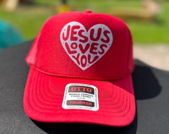 May include: Red trucker hat with a white heart that says "Jesus Loves You" on the front. The hat has a foam front and is made by OTTO.
