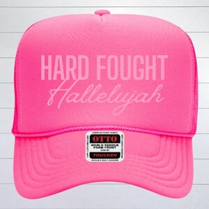 May include: A pink trucker hat with a white mesh back. The front of the hat has the text "HARD FOUGHT Hallelujah" in white.