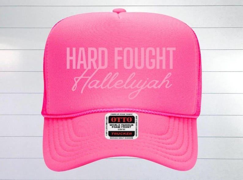 May include: A pink trucker hat with a white mesh back. The front of the hat has the text "HARD FOUGHT Hallelujah" in white.