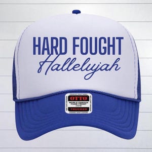 May include: A white trucker hat with a blue mesh back. The front of the hat has the text "HARD FOUGHT Hallelujah" in blue lettering.