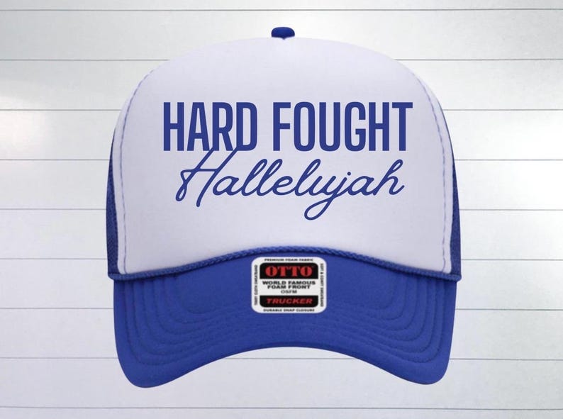 May include: A white trucker hat with a blue mesh back. The front of the hat has the text "HARD FOUGHT Hallelujah" in blue lettering.