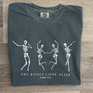 May include: A dark gray t-shirt with a white graphic of three skeletons dancing. The text "Dry Bones Come Alive" and "Ezekiel 37:5" is printed below the skeletons.