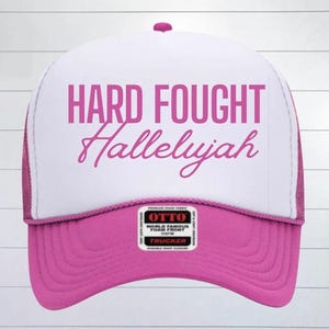 May include: Pink and white trucker hat with the text "HARD FOUGHT Hallelujah" in pink lettering.