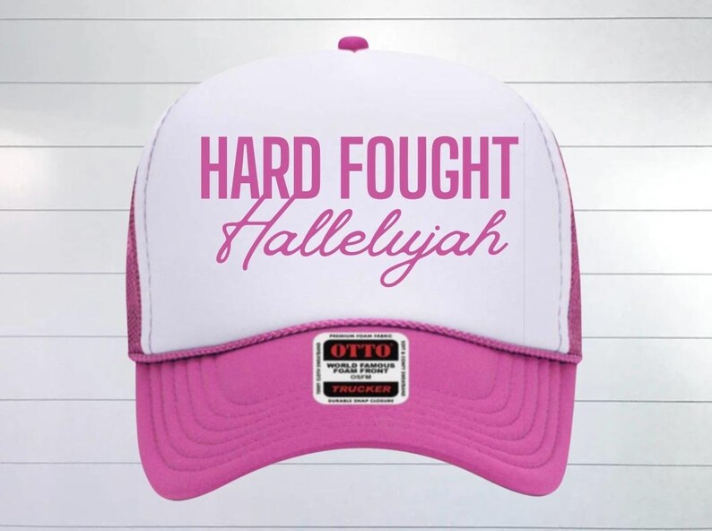 May include: Pink and white trucker hat with the text "HARD FOUGHT Hallelujah" in pink lettering.